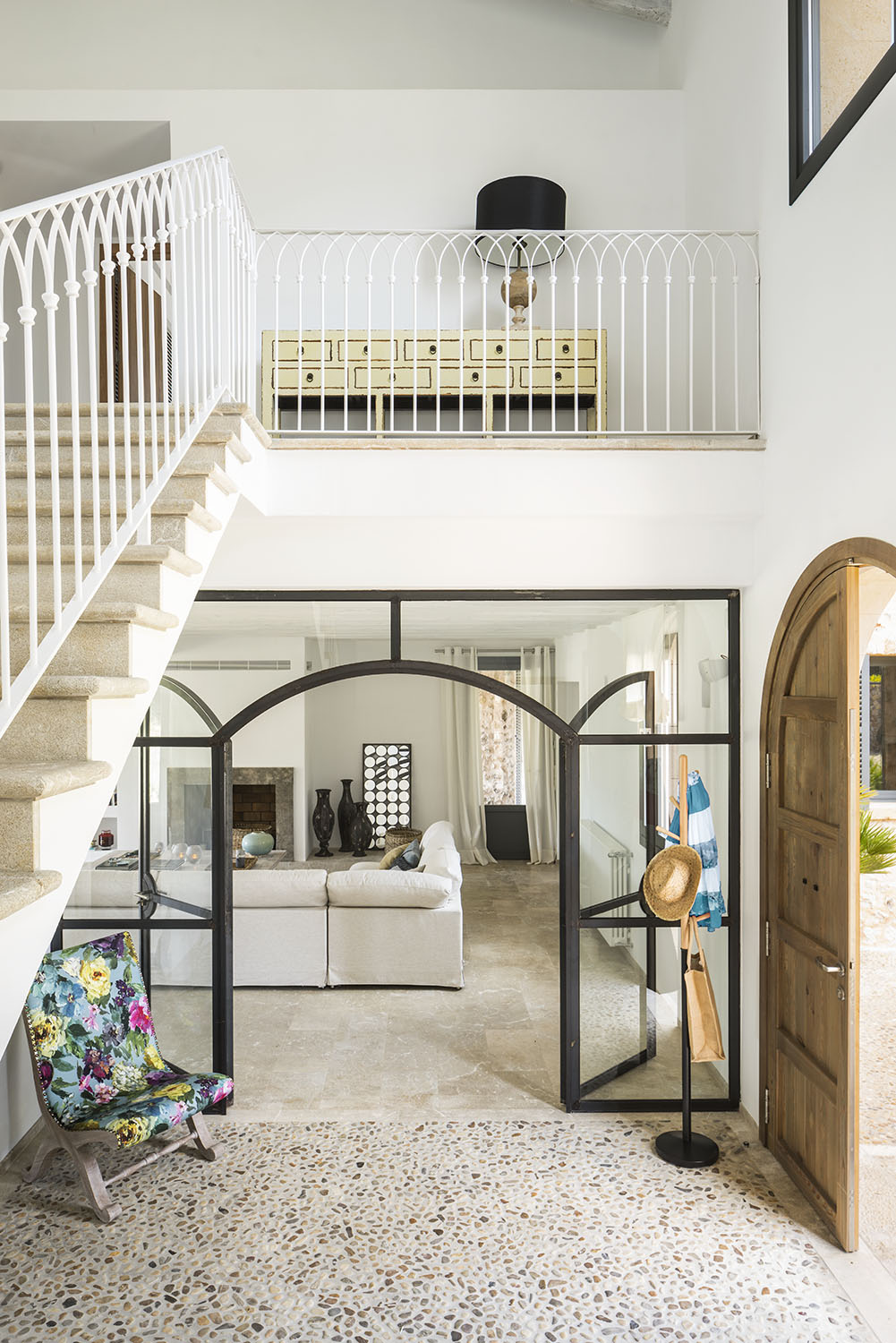 Vestíbulo con escalera de barandilla blanca, cristalera de hierro con arcos, suelo de guijarros y sillón floral