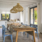 Comedor de la cocina con mesa de madera, sillas azules, dos lámparas de ratán y puerta al jardín