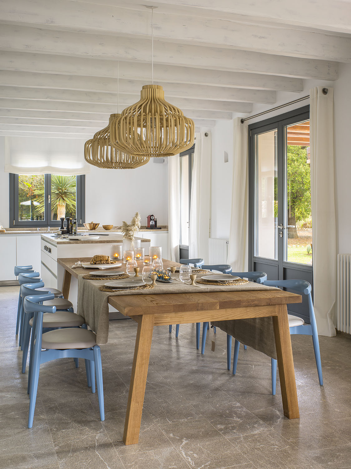 Comedor de la cocina con mesa de madera, sillas azules, dos lámparas de ratán y puerta al jardín