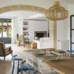 Comedor con mesa de madera puesta, sillas azules, arco de piedra y lámpara de ratán; al fondo, chimenea