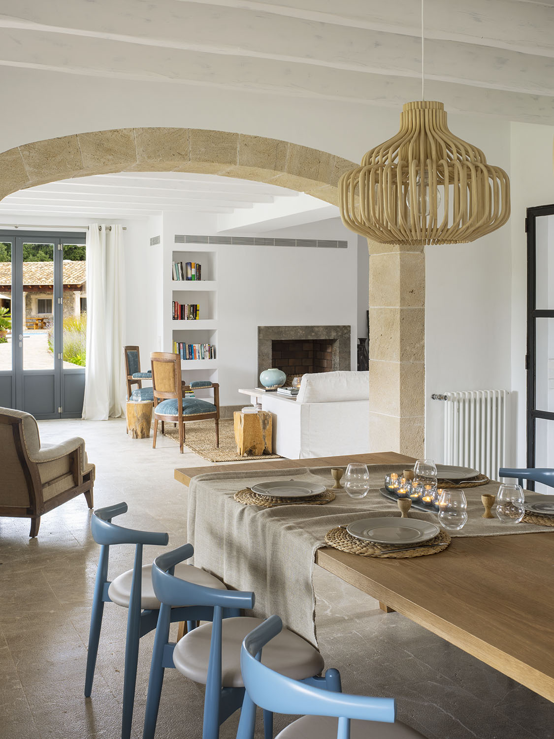 Comedor con mesa de madera puesta, sillas azules, arco de piedra y lámpara de ratán; al fondo, chimenea