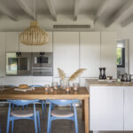 Cocina con armarios blancos de frente liso, hornos empotrados, mesa de madera y sillas azules