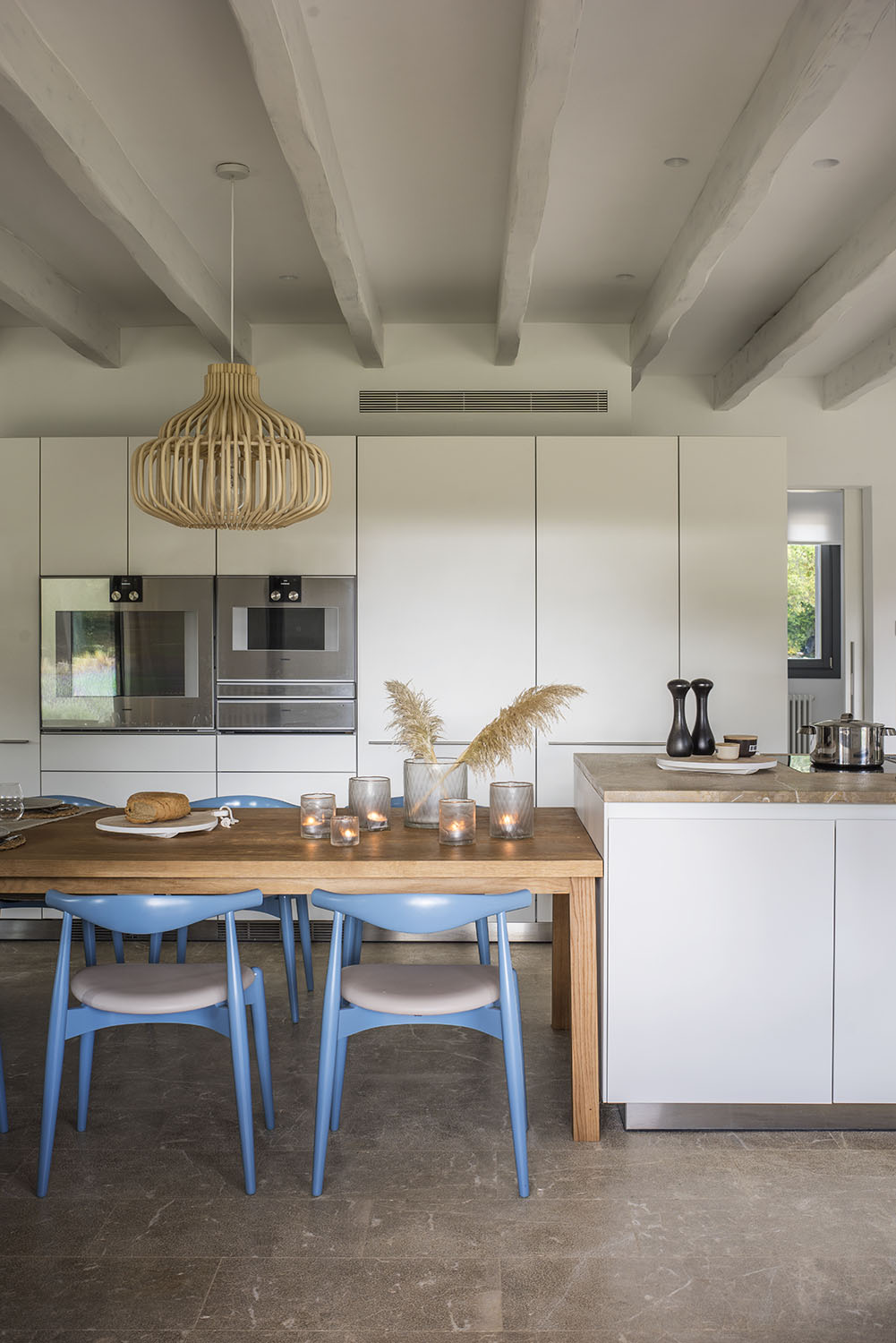 Cocina con armarios blancos de frente liso, hornos empotrados, mesa de madera y sillas azules