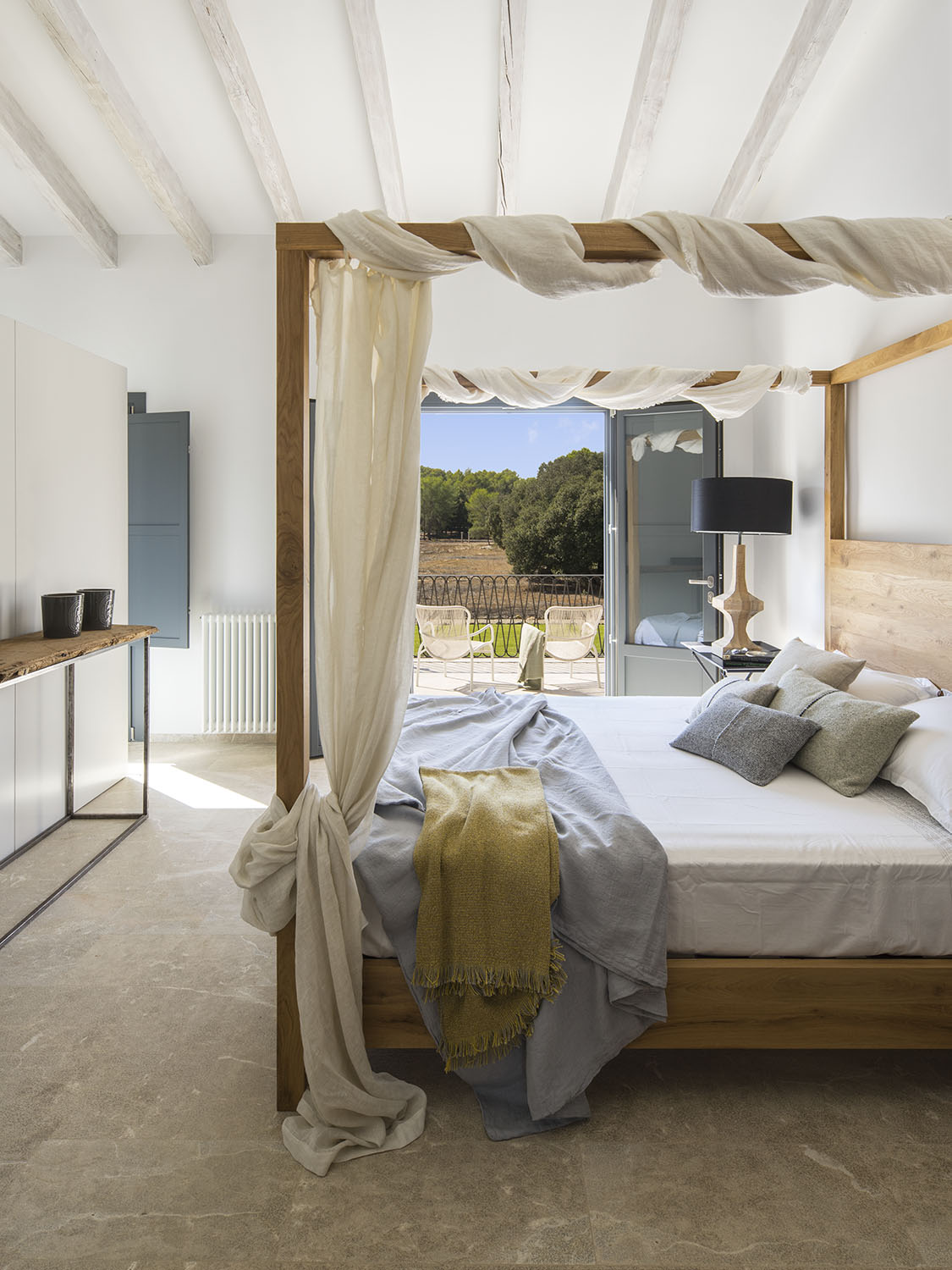 Cama con dosel de madera y gasa, manta amarilla, consola rústica y balcón con sillas blancas