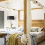 Detalle de la cama de madera con dosel de gasa, manta mostaza y lámpara de pantalla negra