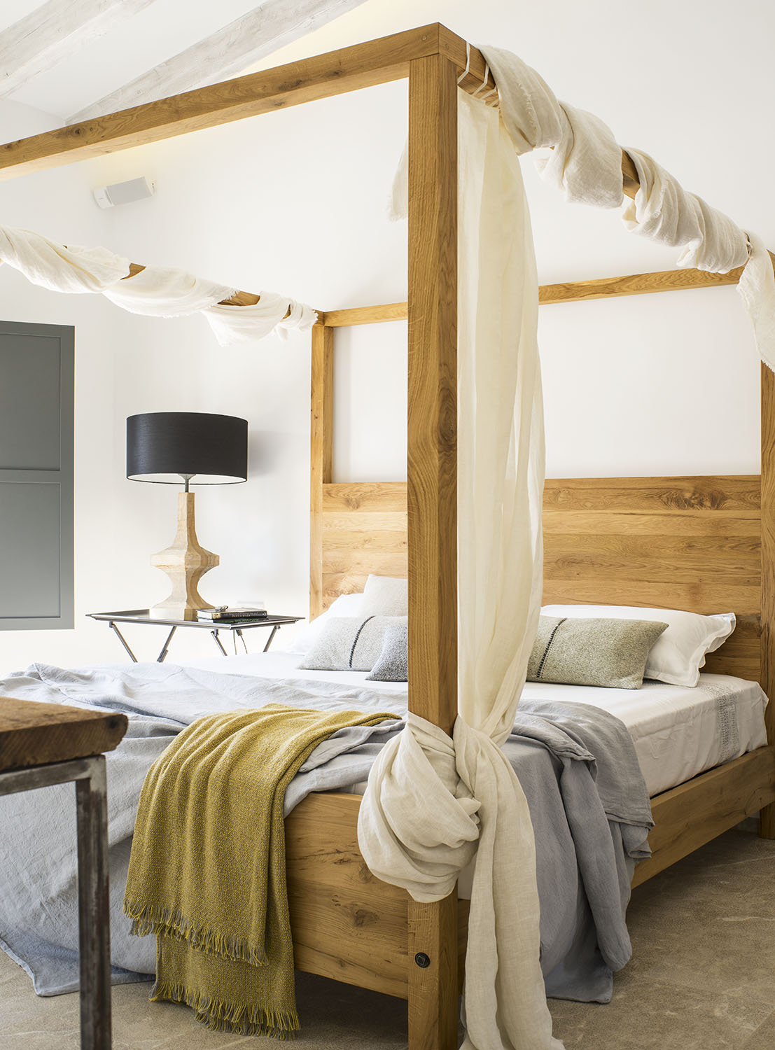 Detalle de la cama de madera con dosel de gasa, manta mostaza y lámpara de pantalla negra