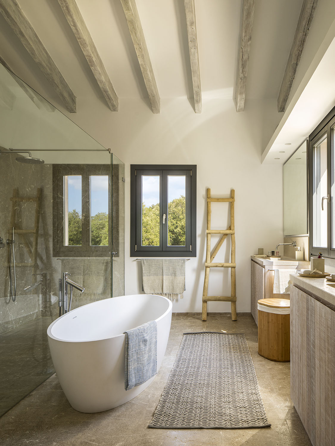 Baño con bañera exenta blanca, ducha acristalada, vigas de madera envejecida y suelo de piedra