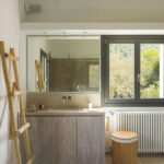 Baño con mueble de lavabo de madera clara, encimera de piedra, espejo amplio y ventana al bosque