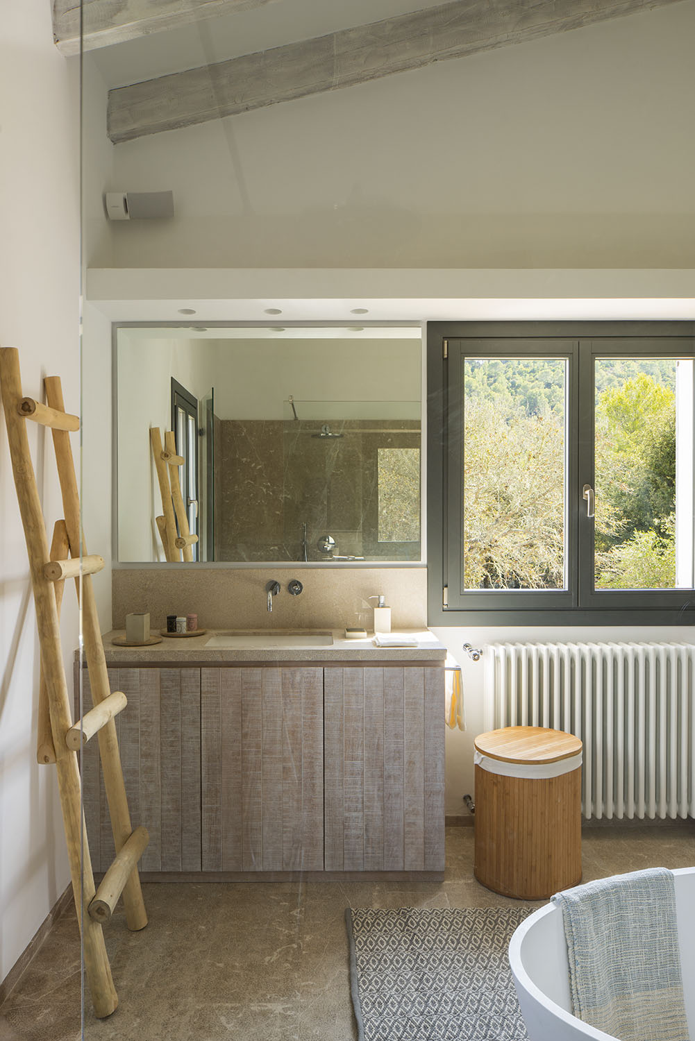 Baño con mueble de lavabo de madera clara, encimera de piedra, espejo amplio y ventana al bosque