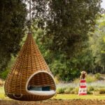 Cama colgante de mimbre en primer plano bajo un árbol, sendero de losas y lavanda al fondo