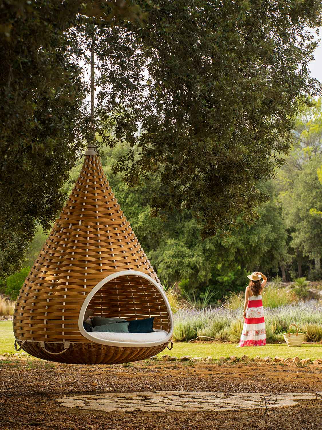 Cama colgante de mimbre en primer plano bajo un árbol, sendero de losas y lavanda al fondo