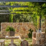 Rincón exterior con pérgola metálica cubierta de enredadera, muro de piedra, mesa y bancos de madera