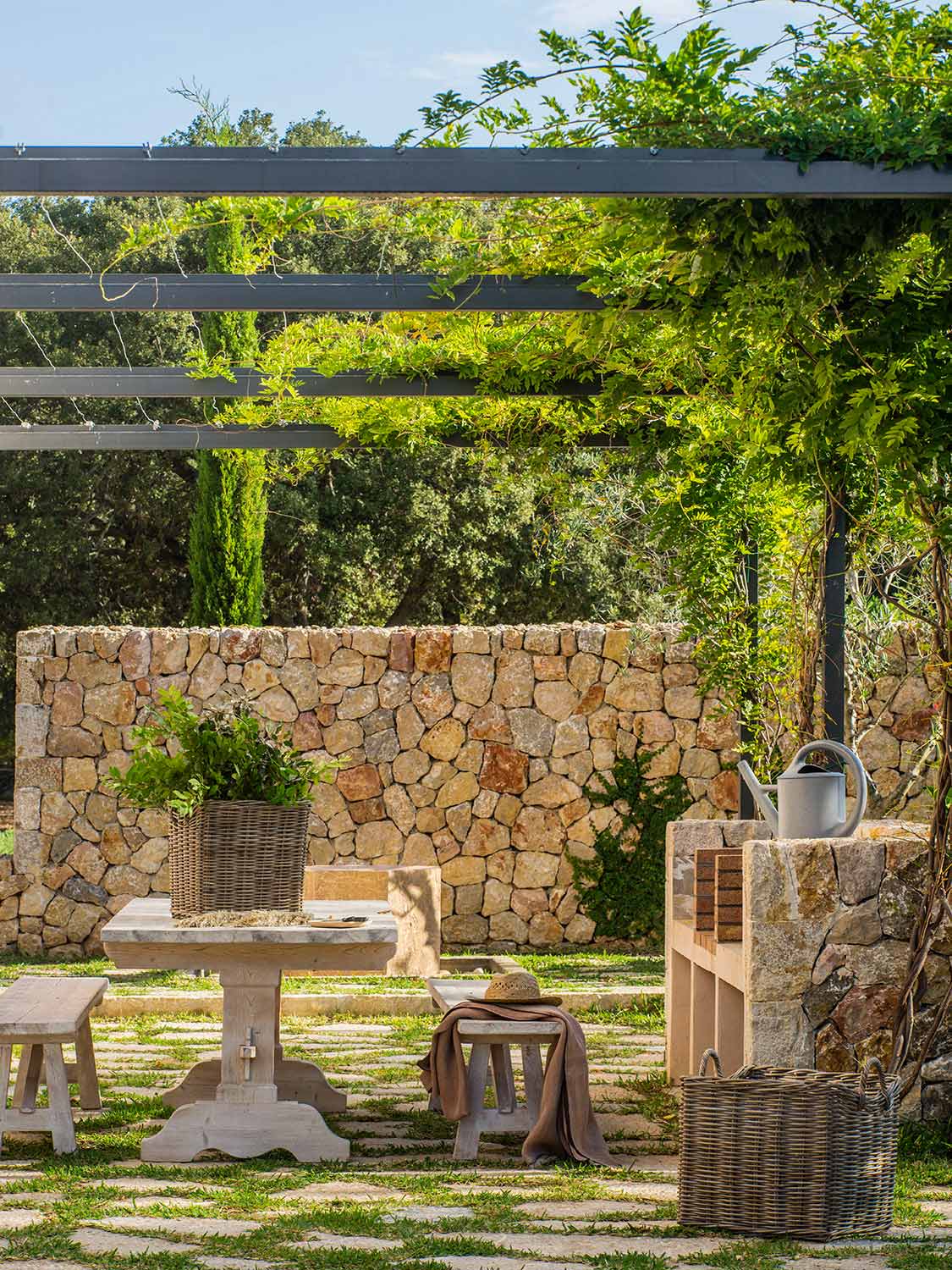 Rincón exterior con pérgola metálica cubierta de enredadera, muro de piedra, mesa y bancos de madera