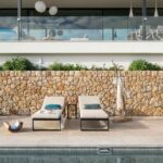 Dos tumbonas beige frente a un muro de piedra junto a la piscina, con terraza acristalada arriba