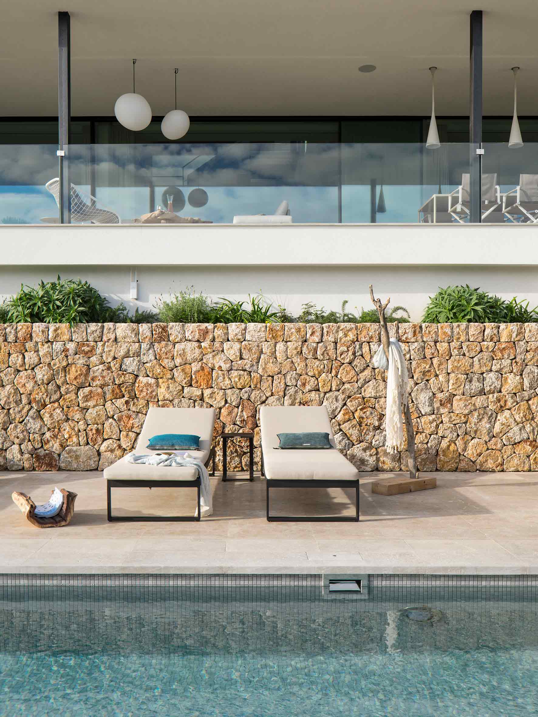Dos tumbonas beige frente a un muro de piedra junto a la piscina, con terraza acristalada arriba