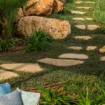 Jardín con camino de losas de piedra sobre el césped, grandes rocas, palmera y cesto de mimbre