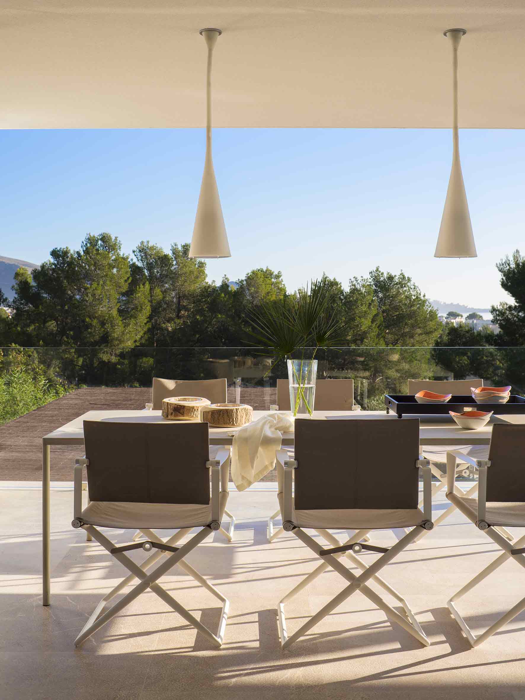 Comedor exterior con sillas plegables, mesa blanca, lámparas colgantes y pinos bajo un cielo azul