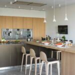 Isla de cocina con barra de madera puesta, taburetes claros, hornos empotrados y lámparas colgantes blancas