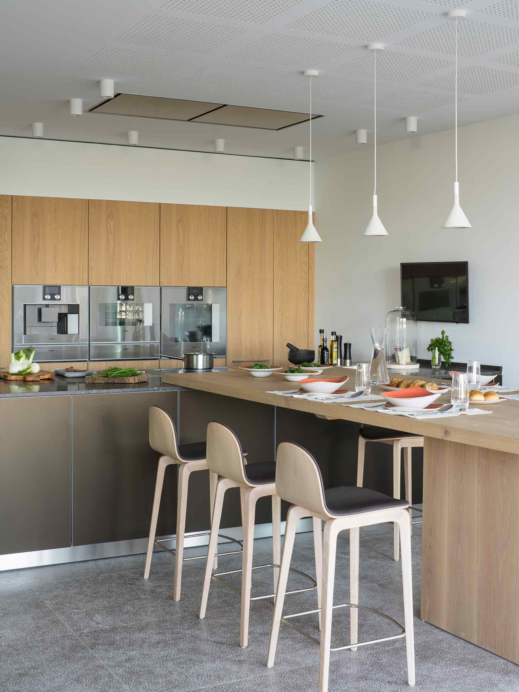 Isla de cocina con barra de madera puesta, taburetes claros, hornos empotrados y lámparas colgantes blancas