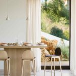 Mesa de madera clara con taburetes junto a un ventanal con rocas y vegetación del jardín