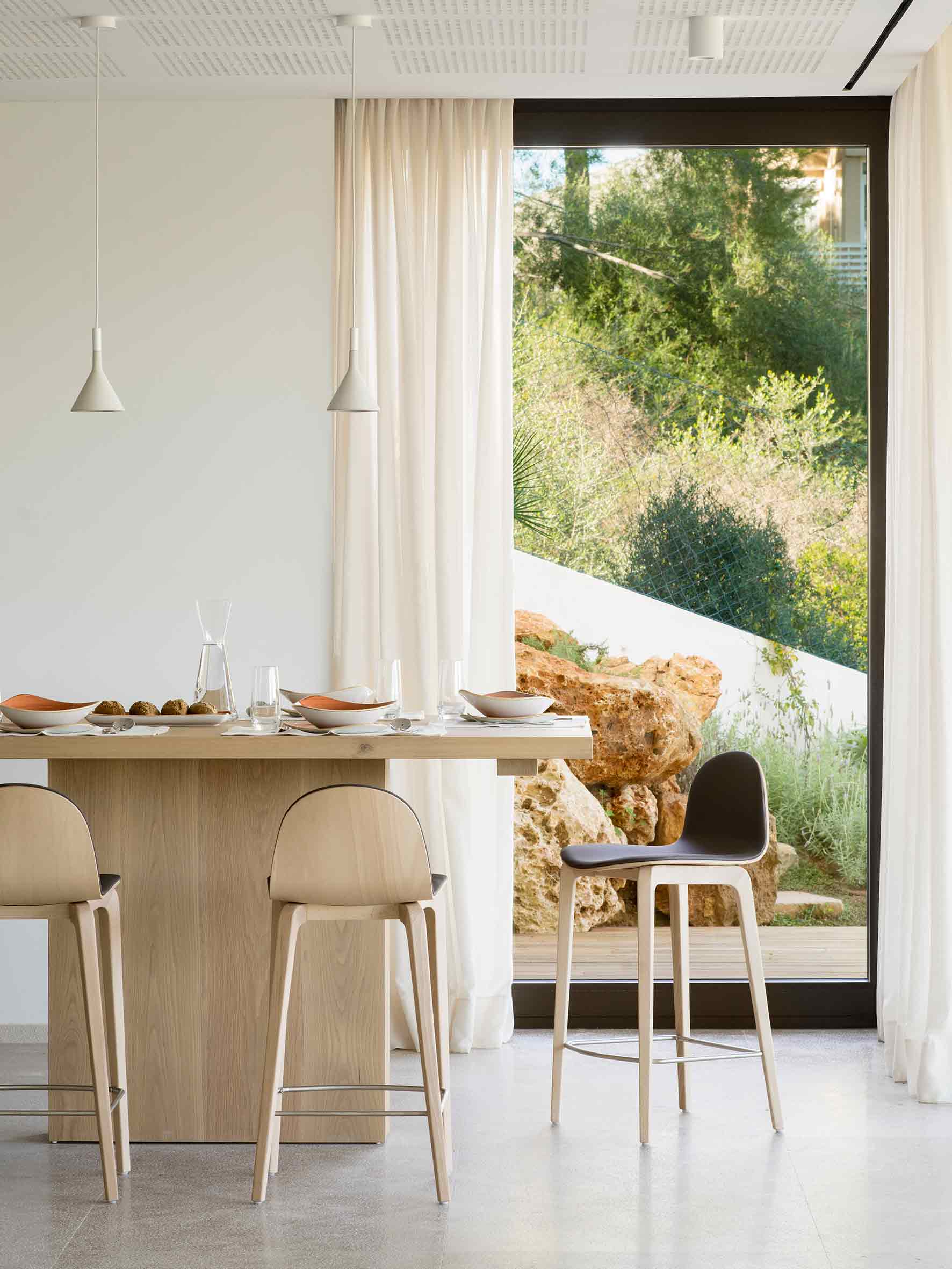Mesa de madera clara con taburetes junto a un ventanal con rocas y vegetación del jardín