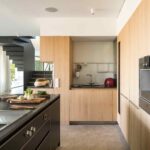 Cocina con isla negra, armarios de roble, fregadero, suelo de piedra gris y escalera al fondo