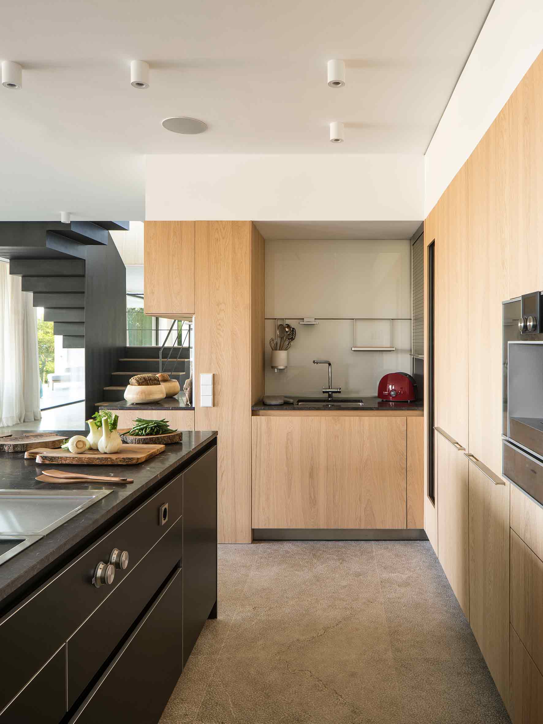 Cocina con isla negra, armarios de roble, fregadero, suelo de piedra gris y escalera al fondo