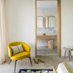 Dormitorio con butaca amarilla y puerta con marco de roble abierta a un baño de mármol blanco