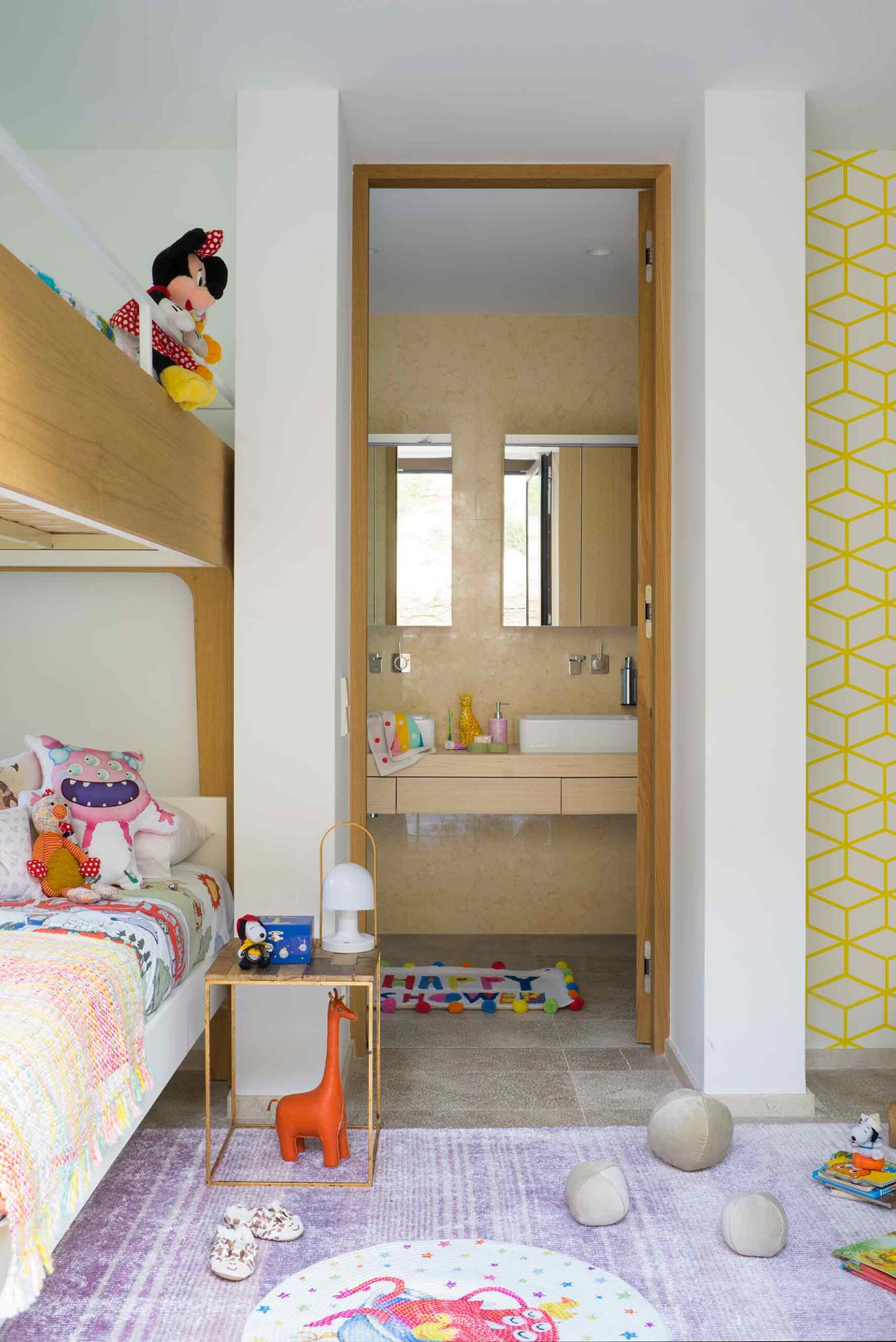 Dormitorio infantil con litera, peluches, papel pintado amarillo y puerta abierta a un baño beige