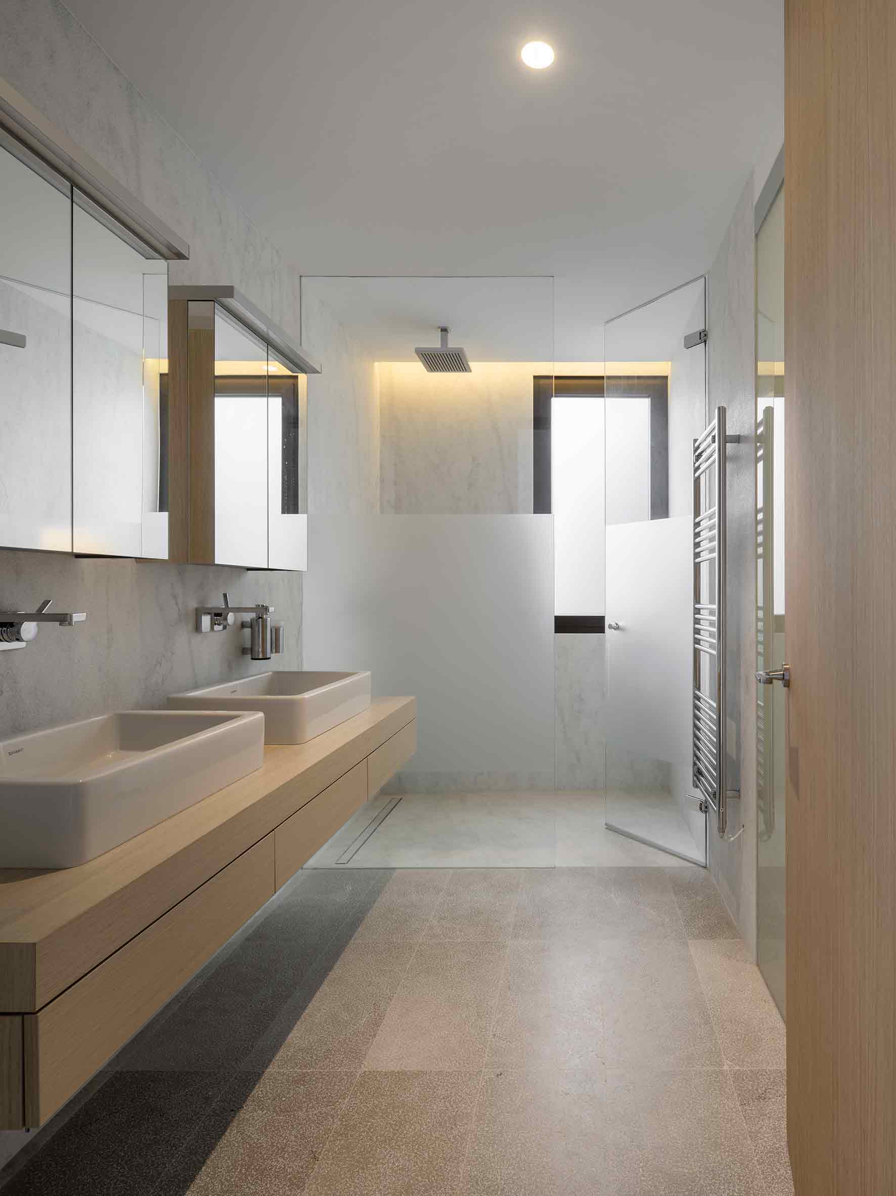 Baño con doble lavabo sobre encimera de roble, ducha de cristal y suelo de piedra clara