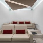 Sala de cine con sofás beige, cojines rojos, dos niveles y paneles de techo con luz indirecta
