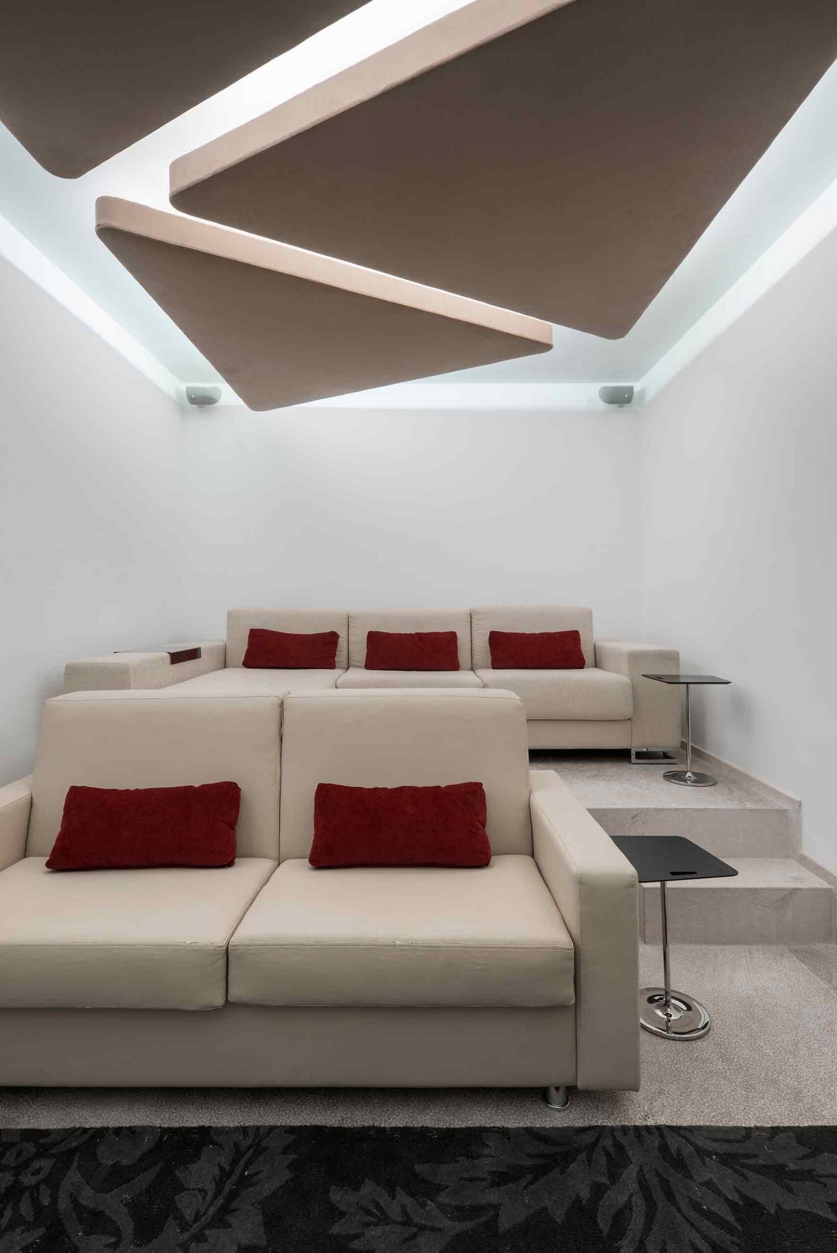 Sala de cine con sofás beige, cojines rojos, dos niveles y paneles de techo con luz indirecta