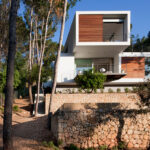 Fachada de la vivienda G138 con lamas de madera, muros de piedra y sendero de tierra entre pinos