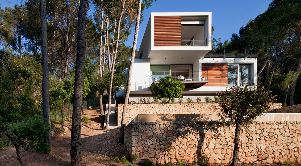 Fachada de la vivienda G138 con lamas de madera, muros de piedra y sendero de tierra entre pinos