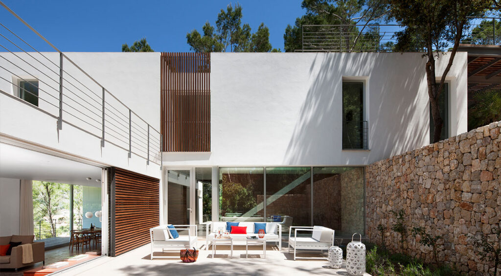 Patio con sofás blancos y cojines de colores frente a cristaleras, celosía de madera y muro de piedra