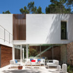 Patio con sofás blancos y cojines de colores frente a cristaleras, celosía de madera y muro de piedra