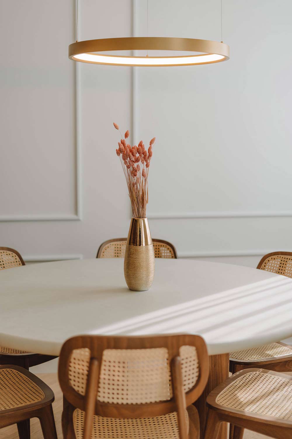Runder Esstisch mit Stühlen aus Rohrgeflecht, goldener Vase mit Trockenblumen und runder Lampe