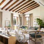 Salón de techo inclinado con vigas de madera, butaca de mimbre, puf trenzado y ventanales al jardín