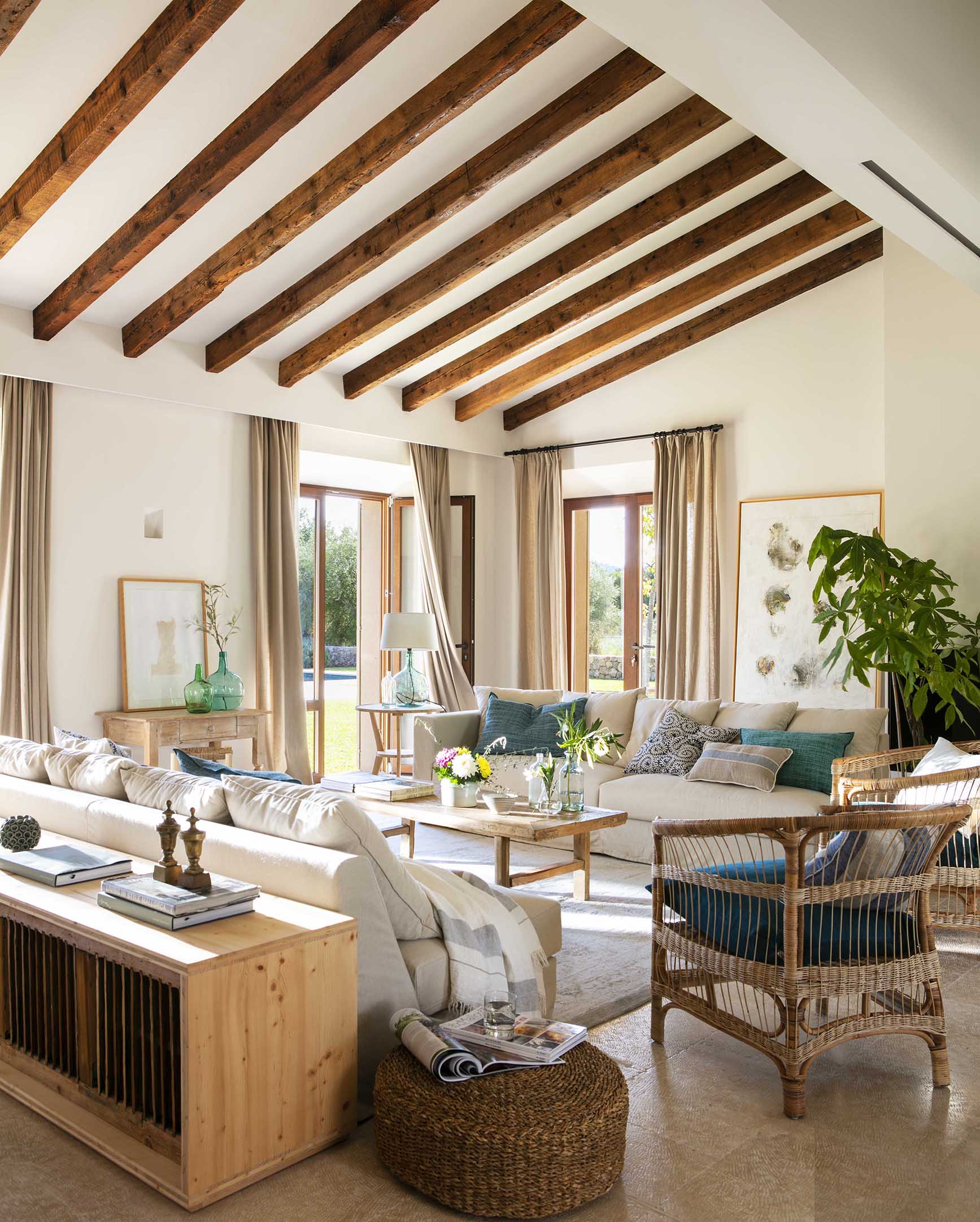 Salón de techo inclinado con vigas de madera, butaca de mimbre, puf trenzado y ventanales al jardín