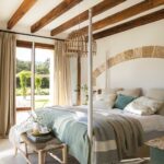 Dormitorio luminoso con cama de cuatro postes, vigas vistas, colcha verde y cristalera hacia el jardín