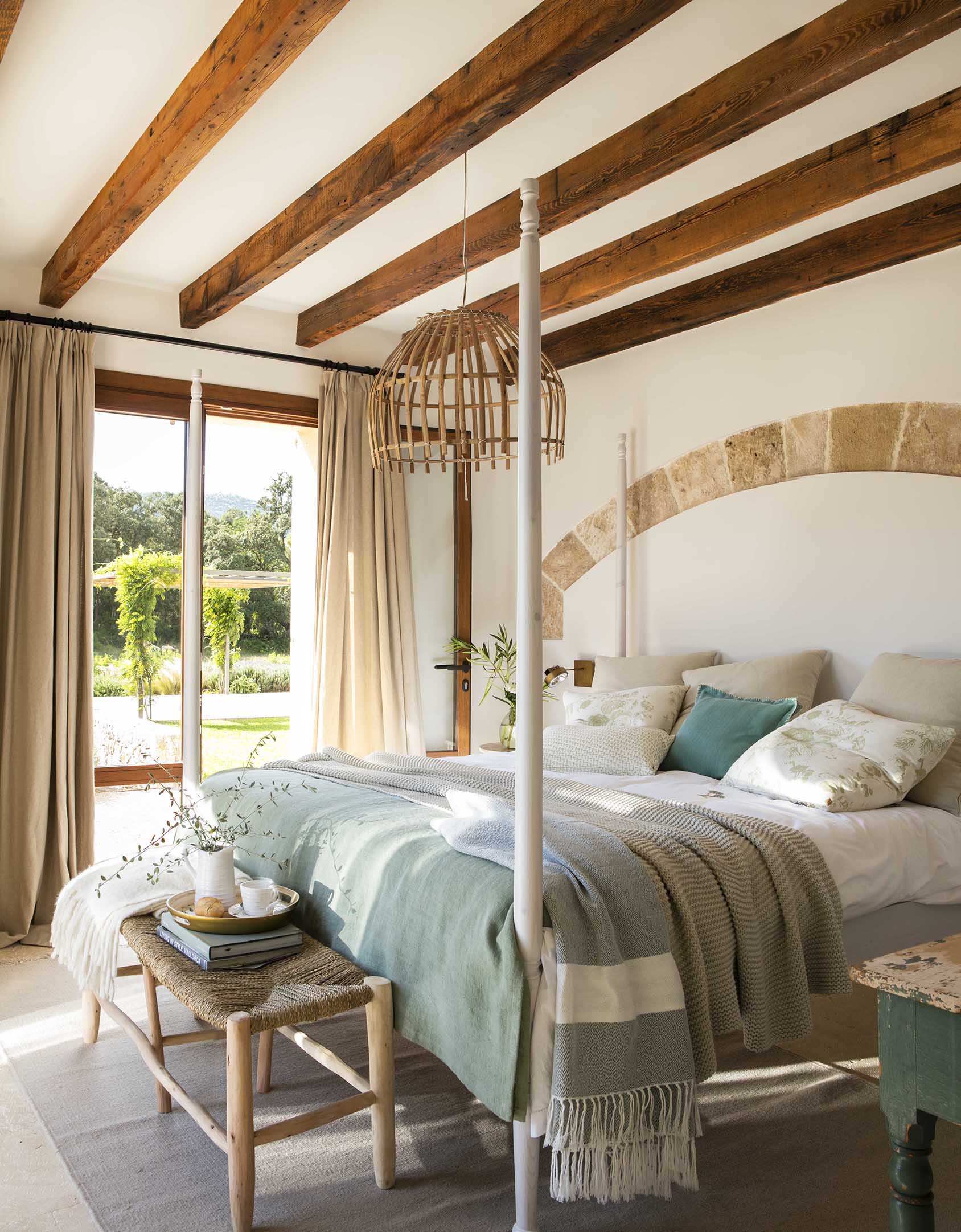 Dormitorio luminoso con cama de cuatro postes, vigas vistas, colcha verde y cristalera hacia el jardín