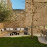 Jardín de Can Angel con sofá de obra, muro de piedra, olivo, césped y comedor exterior