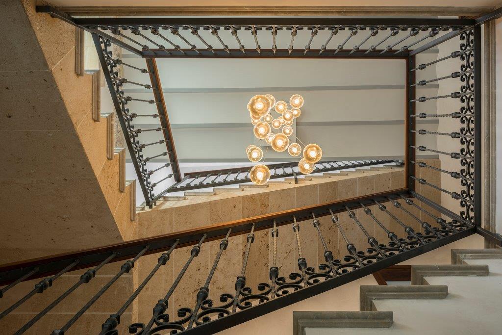 Escalera vista desde abajo con barandilla de forja, piedra clara y lámpara de esferas de cristal