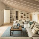 Salón abuhardillado con vigas de madera clara, sofá esquinero, alfombra azul y estanterías blancas empotradas