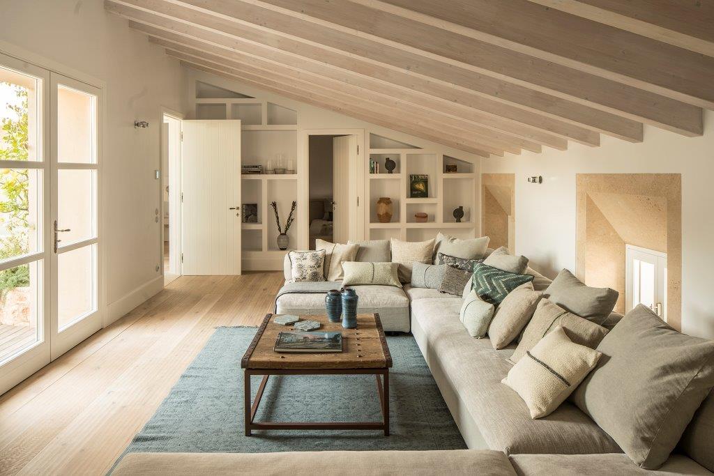 Salón abuhardillado con vigas de madera clara, sofá esquinero, alfombra azul y estanterías blancas empotradas