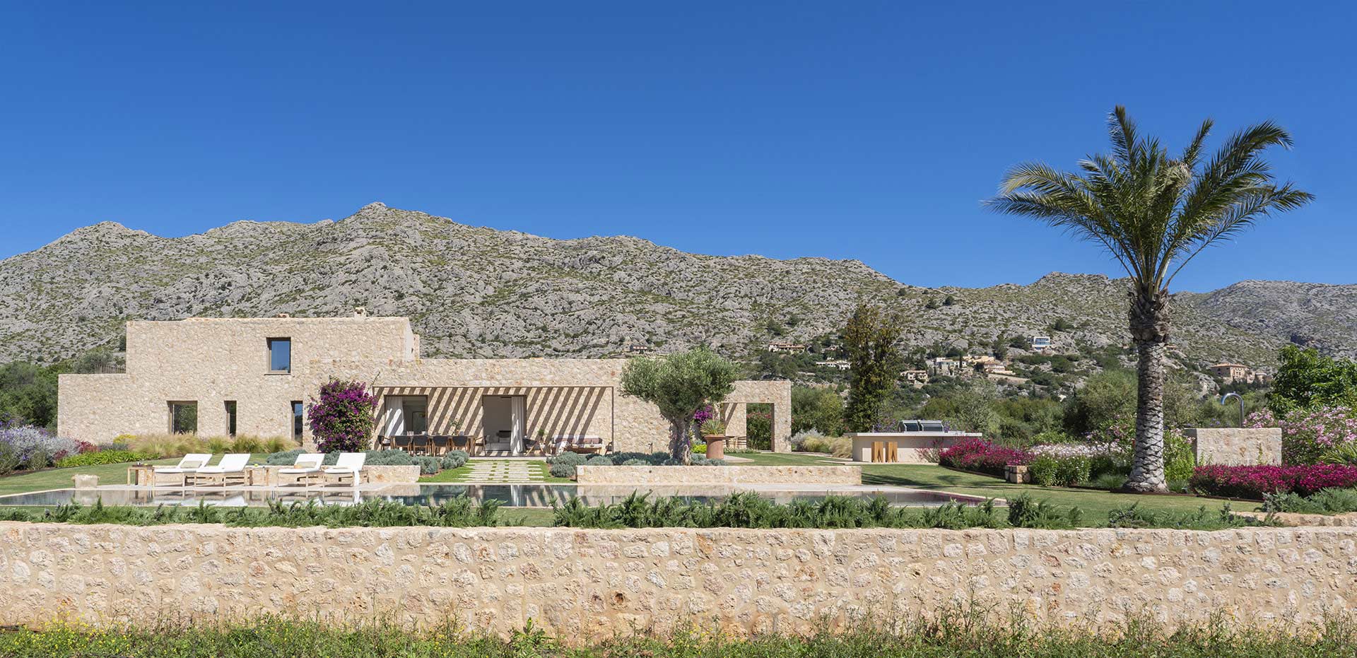 Muro bajo de piedra en primer plano, con la villa, una palmera y la montaña al fondo