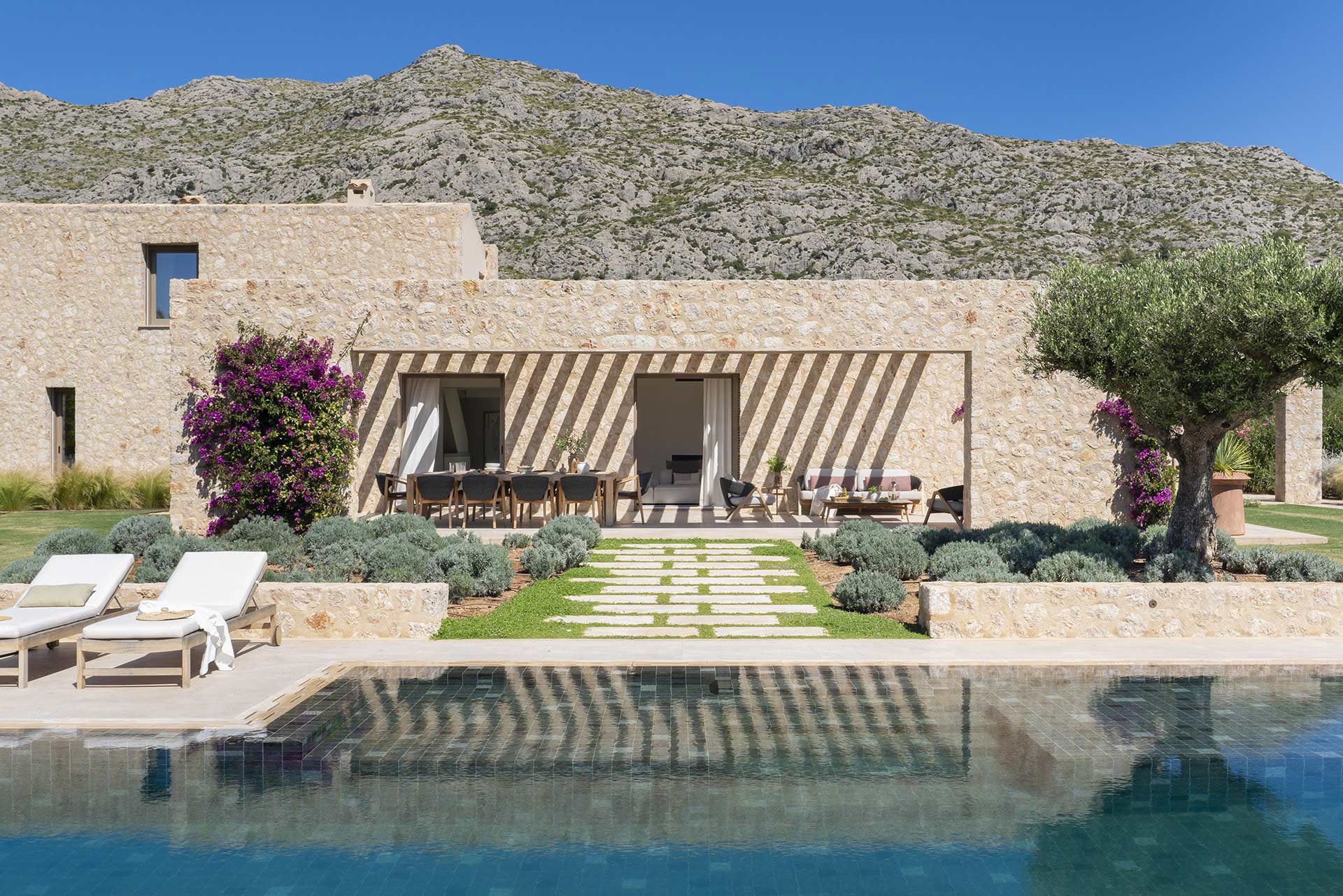 Fachada de piedra de la villa junto a la piscina, con tumbonas, olivo y montaña detrás