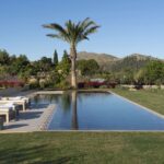 Piscina alargada con tumbonas, palmera y zona de barbacoa bajo luz cálida y montañas al fondo