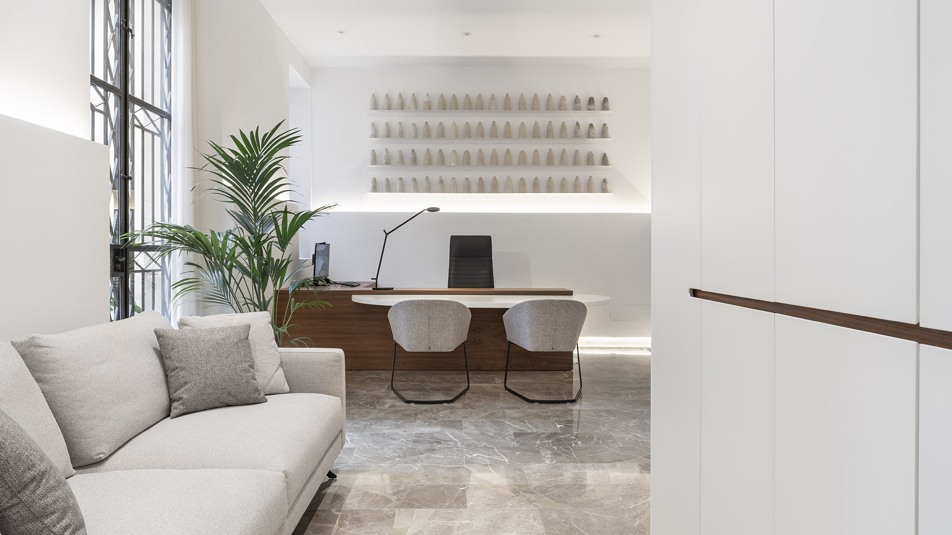 Büro vom hellgrauen Sofa aus gesehen, mit Holzschreibtisch, gepolsterten Stühlen, Palme und weißen Schränken