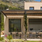 Schilfrohr-Pergola und Putzfassade mit sichtbarer weißer Küche, dahinter ein Berghang
